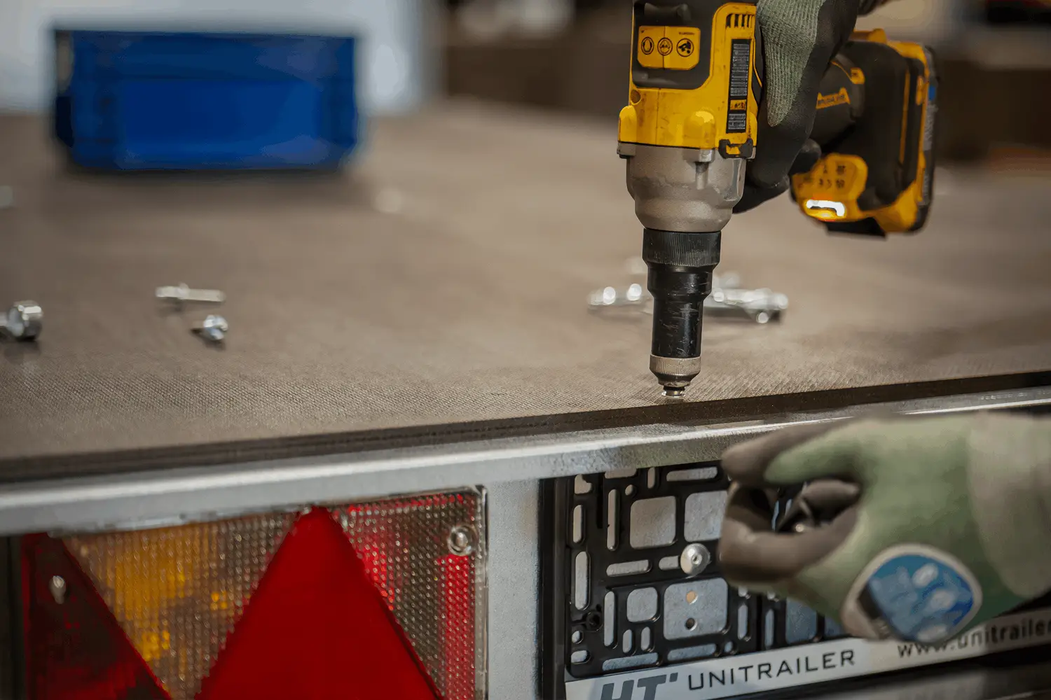 Close-up van de handen van een persoon met werkhandschoenen die een accuboormachine gebruiken om schroeven vast te draaien in de houten vloer van een aanhangwagen.