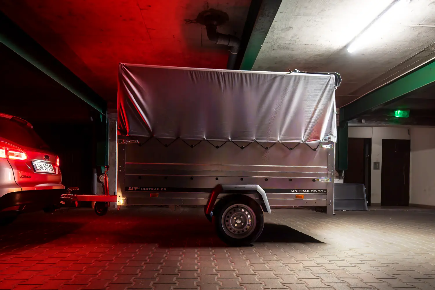 Unitrailer-vrachtaanhangwagen met grijze huif, gekoppeld aan een auto in een slecht verlichte ondergrondse garage.