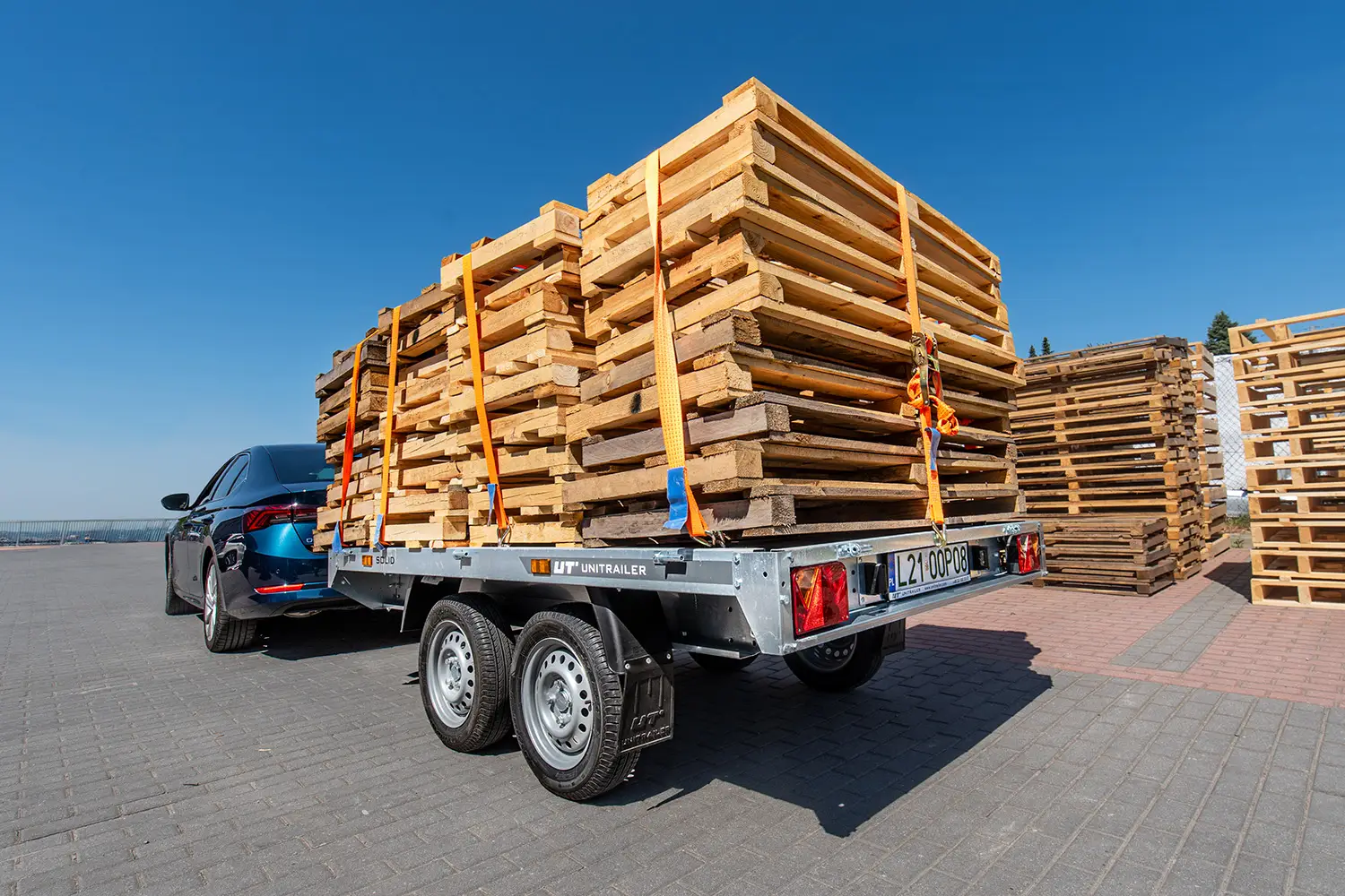 Tandemasser aanhanger van Unitrailer gekoppeld achter een donkere auto, geladen met een hoge stapel houten pallets die zijn vastgezet met spanbanden.