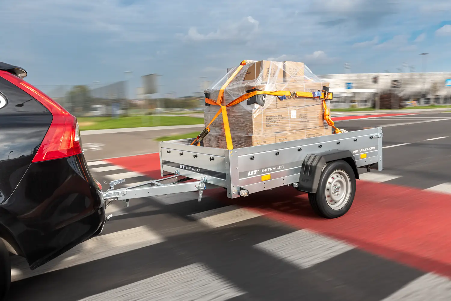 Unitrailer aanhanger gekoppeld achter een zwarte auto, die tijdens het rijden in folie gewikkelde dozen transporteert die zijn vastgezet met spanbanden.