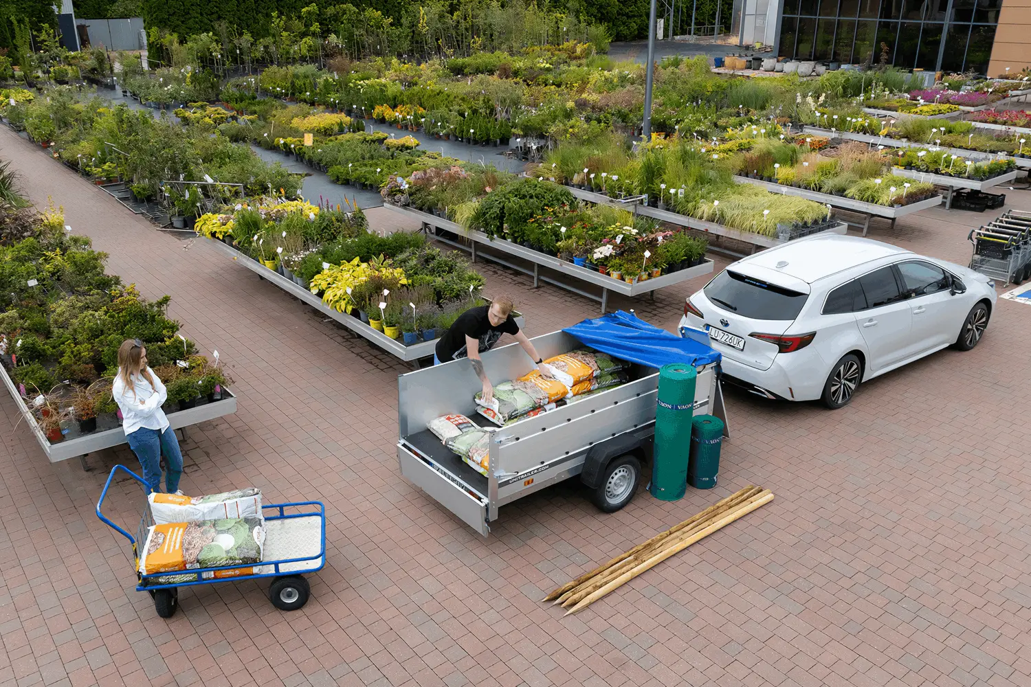 Bovenaanzicht van een tuincentrum waar een witte auto met Unitrailer aanhanger wordt beladen met zakken potgrond en tuinartikelen.